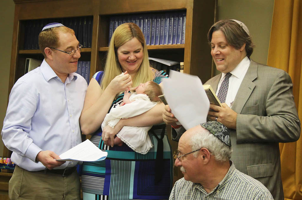 On the left stands a man and woman holding their newborn boy. On the right stands the rabbi reading from a sheath of papers. An older man sits in front of them.