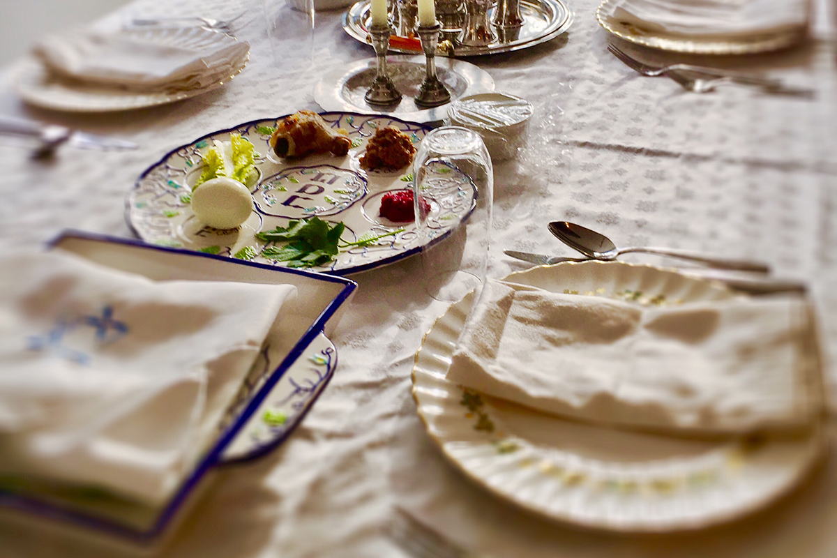 This is a color photo of a table with a white tablecloth, a white and blue seder plate, a plate with a napkin and a plate holding matzah covered with a napkin. There are salt and pepper shakers and part of two candles in the background.