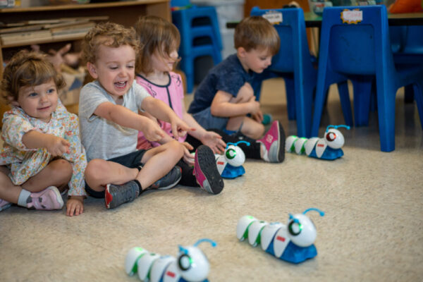 Four children sit in a row on the floor. The first and second children from the left point excitedly at their robots during STEM class.