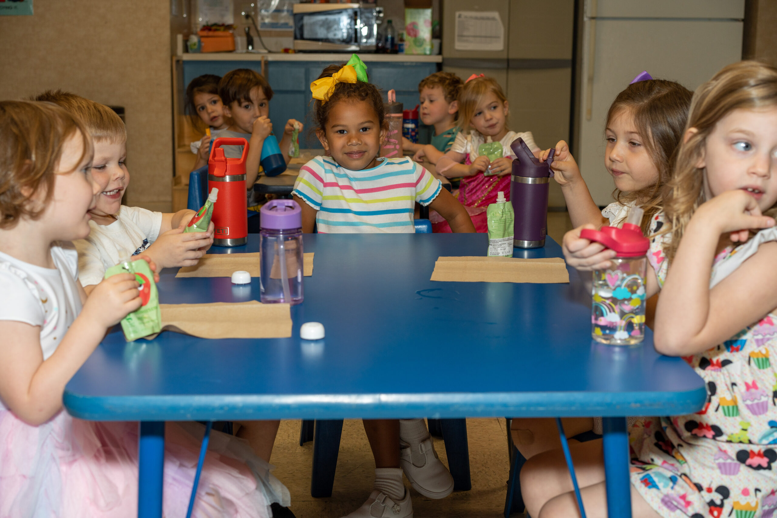 Five children sit at a blue table eating applesauce pouches and drinking water. In the center, a little girl wearing a striped shirt and yellow bow looks at the camera. In the background, four more children sit at another table eating.