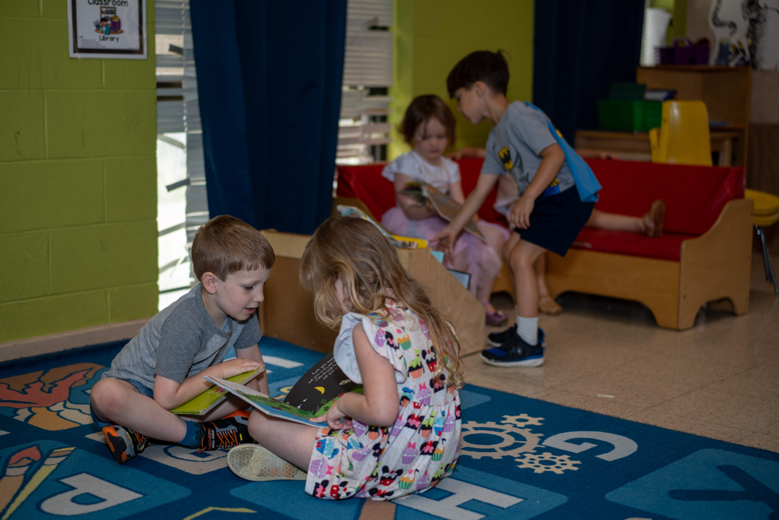 In the foreground, two preschoolers sit on a rug looking at a book. In the background, another preschooler looks at a book while a fourth child reaches for a book on a shelf.