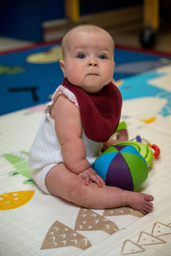 A bald baby with big eyes and big cheeks wearing a white onesie and a red bib sits on the floor, looking directly at the camera with several toys between her legs