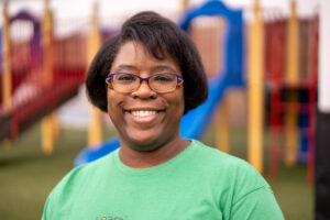 A friendly looking woman with a large smile looks at the camera. She is wearing a green shirt and standing in front of playground equipment