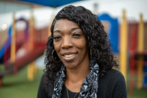A woman smiles warmly at the camera. She is wearing a black shirt, black and white scarf and gold hoop earrings. She is standing in front of playground equipment