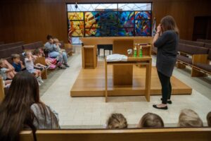 Rabbi Sarah stands in front of the bimah holding her hands over her eyes as she pretends to light play Shabbat candles. Several children sitting on a pew to the left mimic her.
