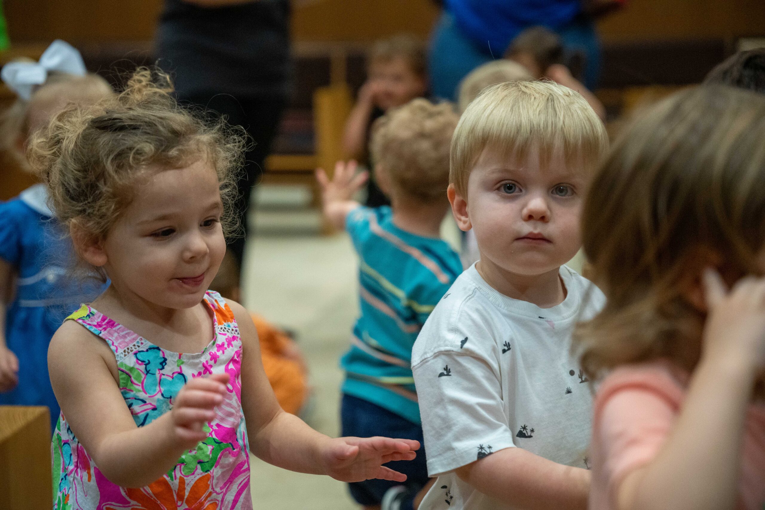 This photo is taken at toddler eye level. One boy in a white shirt with blond hair looks at the camera. A little girl with curly hair and a multi-colored shirt looks to the side. More children are in the background out of focus.