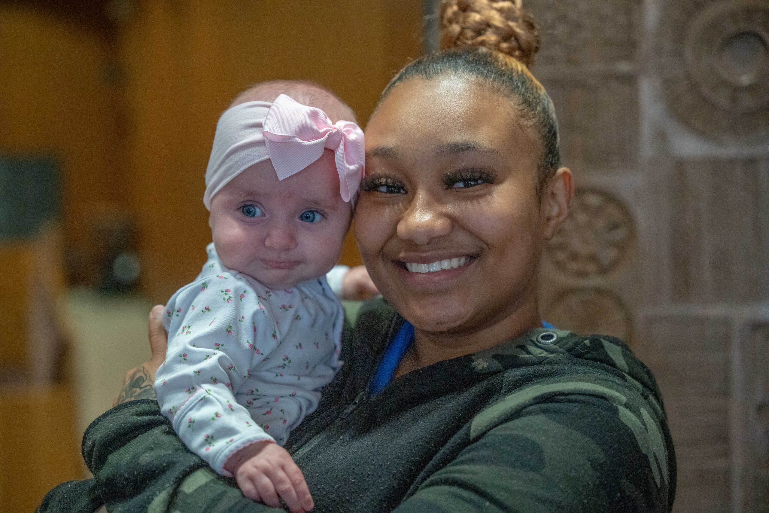 A young woman in a sweatshirt with a big smile holds a baby girl in a white onesie and large pink bow.