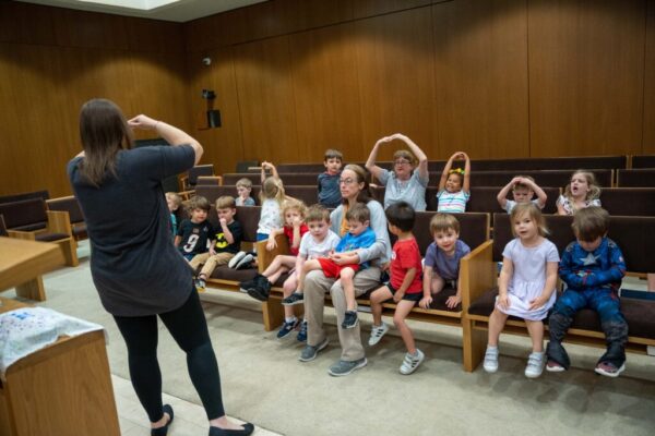 Rabbi Sarah stands in front of children and teachers seated in a pew directing them in a song. Rabbi and several children have their hands above their heads in a circle